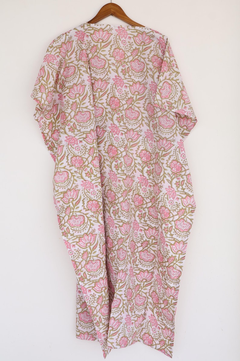 May include: A white and pink floral print kaftan dress hanging on a wooden hanger. The dress features a repeating pattern of pink flowers and green leaves. The kaftan is made of a lightweight fabric.