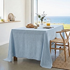 May include: A light blue linen tablecloth covers a wooden table set for a meal. A wooden chair with a woven seat sits nearby. The table is set with plates, bowls, and a basket of bread. A vase of flowers and a pitcher of juice are also on the table.