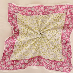 May include: A square, folded textile with a floral pattern. The center features a white background with small green and gray floral sprigs. A vibrant pink border with white floral designs frames the center. The textile is likely cotton.