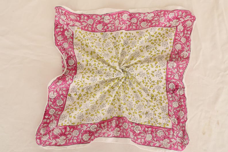 May include: A square, folded textile with a floral pattern. The center features a white background with small green and gray floral sprigs. A vibrant pink border with white floral designs frames the center. The textile is likely cotton.