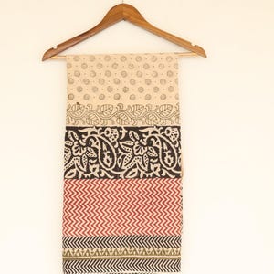 May include: A beige textile with a repeating dot pattern, floral and geometric designs in black, red, and green. The fabric is displayed on a wooden hanger against a white background. The textile is likely a scarf or a piece of fabric.