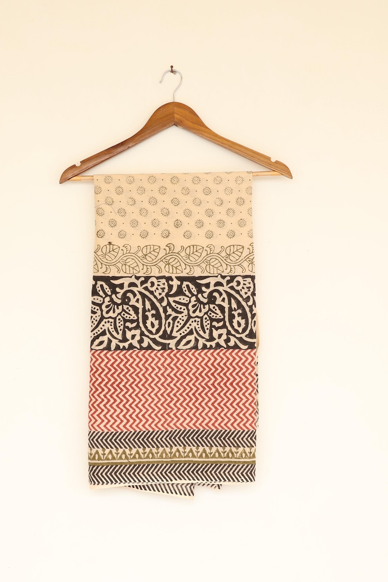 May include: A beige textile with a repeating dot pattern, floral and geometric designs in black, red, and green. The fabric is displayed on a wooden hanger against a white background. The textile is likely a scarf or a piece of fabric.