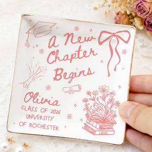 May include: A white square ceramic trinket dish with gold trim. The dish features pink illustrations and text that reads "a New Chapter Begins" and "Olivia Class of 2026 University of Rochester". Two pearl earrings are in the foreground.