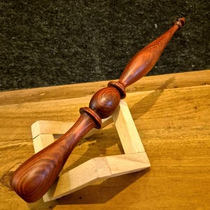 May include: A finely crafted wooden wand, displaying a deep reddish-brown colour. The wand has a rounded central section and a slender handle and tip. It rests on a small, light-coloured wooden stand, highlighting the detailed wood grain and artistry.