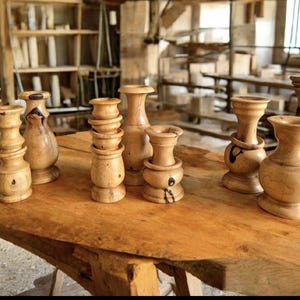 May include: A selection of turned wooden vases and decorative items in different shapes and sizes. They are arranged on a large, roughly-hewn wooden table, highlighting the natural wood grain. The background suggests a workshop or studio environment.