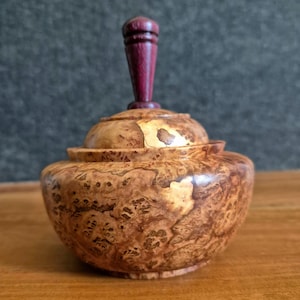 May include: A small, round wooden lidded container featuring a polished, swirling brown burl wood grain pattern. The lid is topped with a smooth, vertical, dark purple wooden finial handle that contrasts with the natural tan and brown tones.