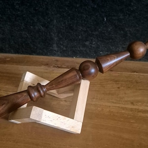 May include: A dark brown wooden wand with a series of rounded and conical shapes. The wand is displayed on a light-coloured wooden stand. The background is dark, with the wand's details clearly visible.