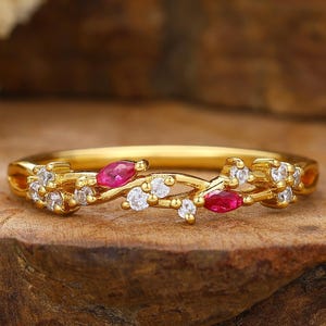 May include: A gold ring with a delicate design featuring small, round white gemstones and oval-shaped red gemstones. The ring is set against a natural, textured background, highlighting its intricate details.