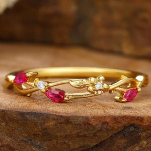May include: A gold ring with a floral design. The ring features three oval-cut, bright red gemstones and small, round, clear gemstones. The band has a delicate, vine-like pattern. The ring is set on a wooden surface.