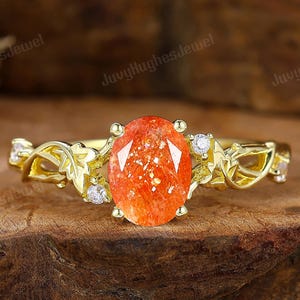 May include: A gold ring with an oval, orange gemstone center. The ring features a floral design with small, clear accent stones. The ring is set on a wooden surface.