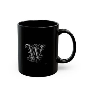 May include: A solid black ceramic mug featuring a white, ornate, calligraphic letter W on the front. The letter is designed with intricate, swirling, vine-like flourishes and sharp, pointed edges, creating a detailed, high-contrast graphic.