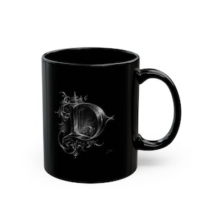 May include: A glossy black ceramic mug featuring a large, intricate, silver-toned letter D on the front. The letter is rendered in a detailed, ornate calligraphic style with swirling flourishes and decorative elements surrounding the character.