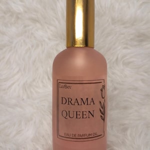 May include: A pink perfume oil bottle with a gold cap. The label reads "Drama Queen" with "Eau de Parfum Oil" below. The bottle has a frosted appearance and is set against a fluffy white background.