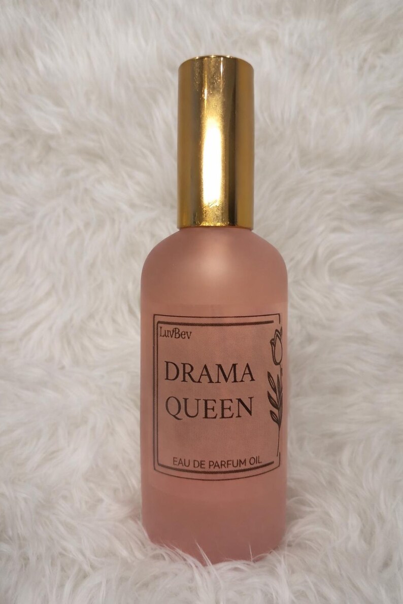 May include: A pink perfume oil bottle with a gold cap. The label reads "Drama Queen" with "Eau de Parfum Oil" below. The bottle has a frosted appearance and is set against a fluffy white background.