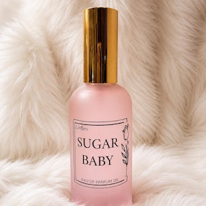 May include: A frosted pink glass bottle of LuvBev Sugar Baby eau de parfum oil with a shiny gold cylindrical cap. The rectangular label features black text reading LuvBev, SUGAR BABY, and EAU DE PARFUM OIL, accompanied by a simple line drawing of a tulip.