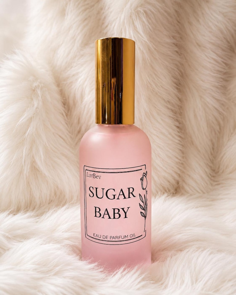 May include: A frosted pink glass bottle of LuvBev Sugar Baby eau de parfum oil with a shiny gold cylindrical cap. The rectangular label features black text reading LuvBev, SUGAR BABY, and EAU DE PARFUM OIL, accompanied by a simple line drawing of a tulip.