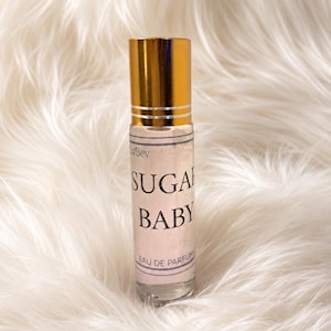May include: A clear glass rollerball perfume bottle with a shiny gold cap. The label features the text "Sugar Baby" in bold black lettering, with "Eau de Parfum" printed below. The bottle contains a light-colored liquid and has a cylindrical shape.