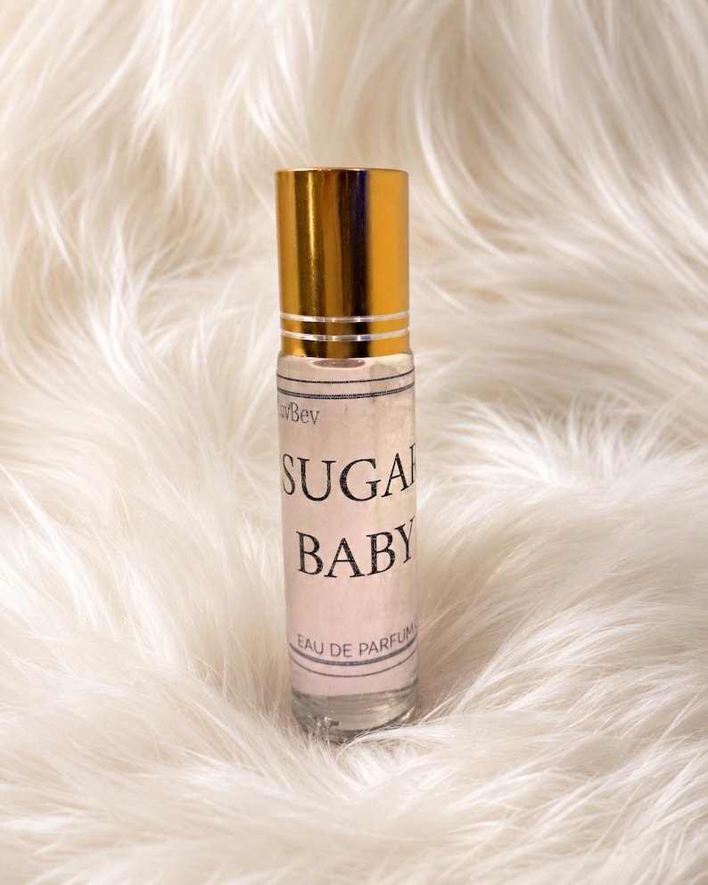 May include: A clear glass rollerball perfume bottle with a shiny gold cap. The label features the text "Sugar Baby" in bold black lettering, with "Eau de Parfum" printed below. The bottle contains a light-colored liquid and has a cylindrical shape.