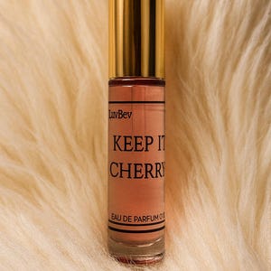 May include: A cylindrical glass perfume roller bottle with a shiny gold cap. The label features the text LuvBev, KEEP IT CHERRY, and EAU DE PARFUM. The liquid inside is a soft peach color, and the bottle is clear glass with black horizontal lines.