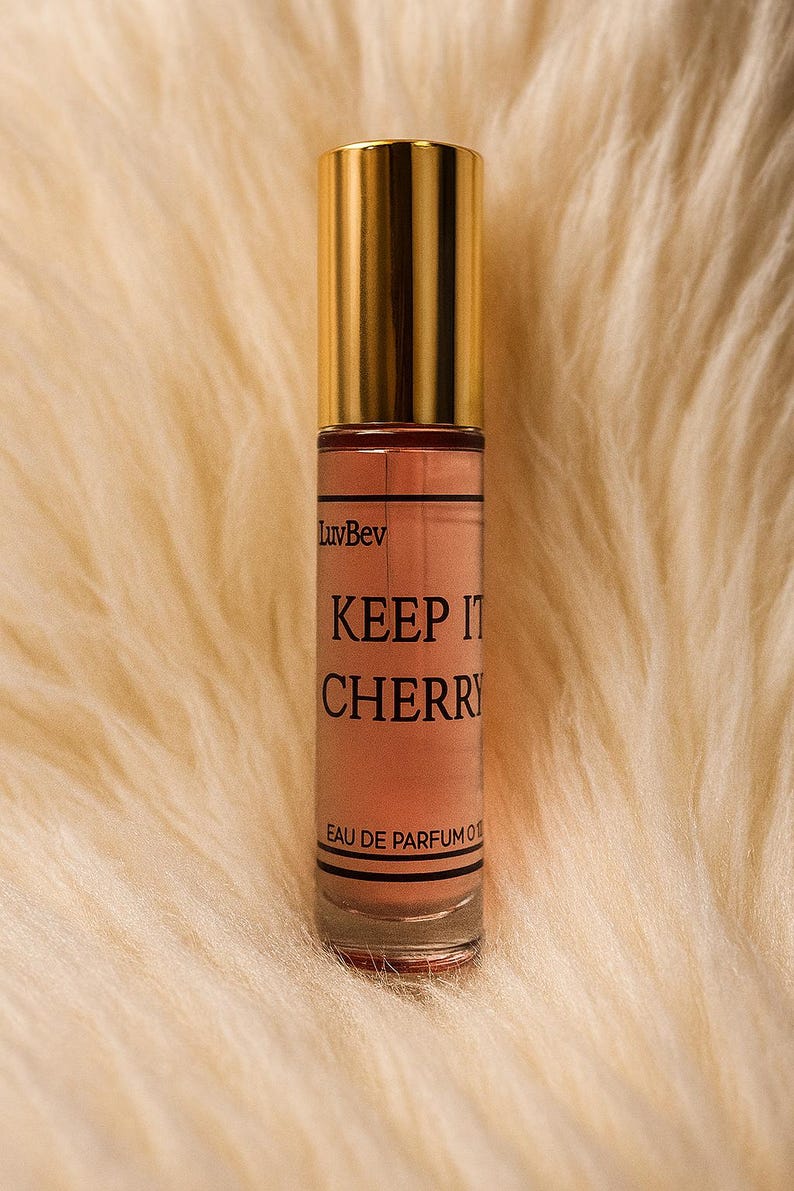 May include: A cylindrical glass perfume roller bottle with a shiny gold cap. The label features the text LuvBev, KEEP IT CHERRY, and EAU DE PARFUM. The liquid inside is a soft peach color, and the bottle is clear glass with black horizontal lines.