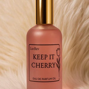 May include: A frosted pink glass perfume bottle with a gold cap. The rectangular label features the text LuvBev, KEEP IT CHERRY, and EAU DE PARFUM OIL, accompanied by a black line drawing of a tulip flower on the right side of the label.