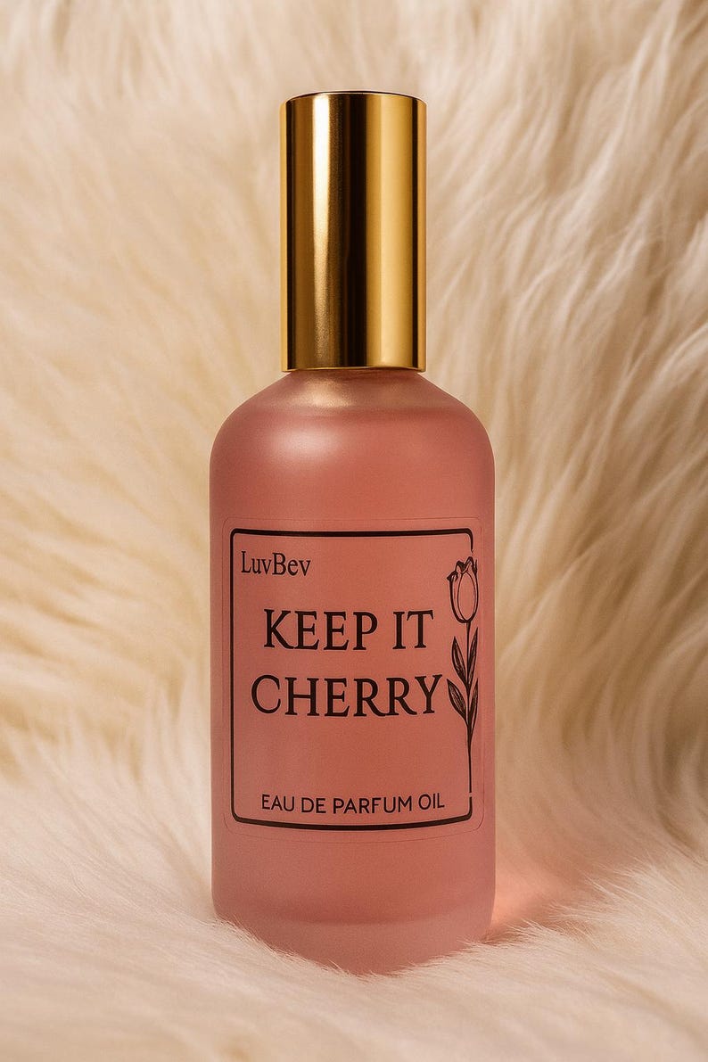 May include: A frosted pink glass perfume bottle with a gold cap. The rectangular label features the text LuvBev, KEEP IT CHERRY, and EAU DE PARFUM OIL, accompanied by a black line drawing of a tulip flower on the right side of the label.