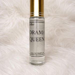 May include: A clear glass perfume bottle with a gold cap. The label reads "DRAMA QUEEN" in black text. The words "Eau de Parfum" are printed at the bottom. The bottle is set against a white, fluffy background.
