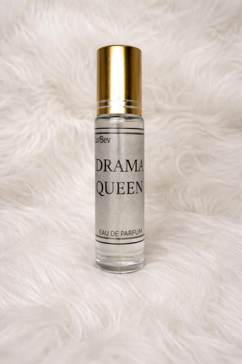 May include: A clear glass perfume bottle with a gold cap. The label reads "DRAMA QUEEN" in black text. The words "Eau de Parfum" are printed at the bottom. The bottle is set against a white, fluffy background.