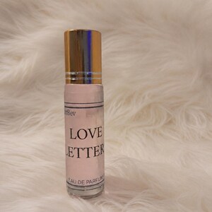 Puede incluir: Un frasco de perfume cil&iacute;ndrico de vidrio con una tapa de metal dorado. La etiqueta es de color rosa claro con texto negro que dice Love Letter y Eau de Parfum. El frasco contiene un l&iacute;quido transparente y tiene un aplicador de bola arriba.