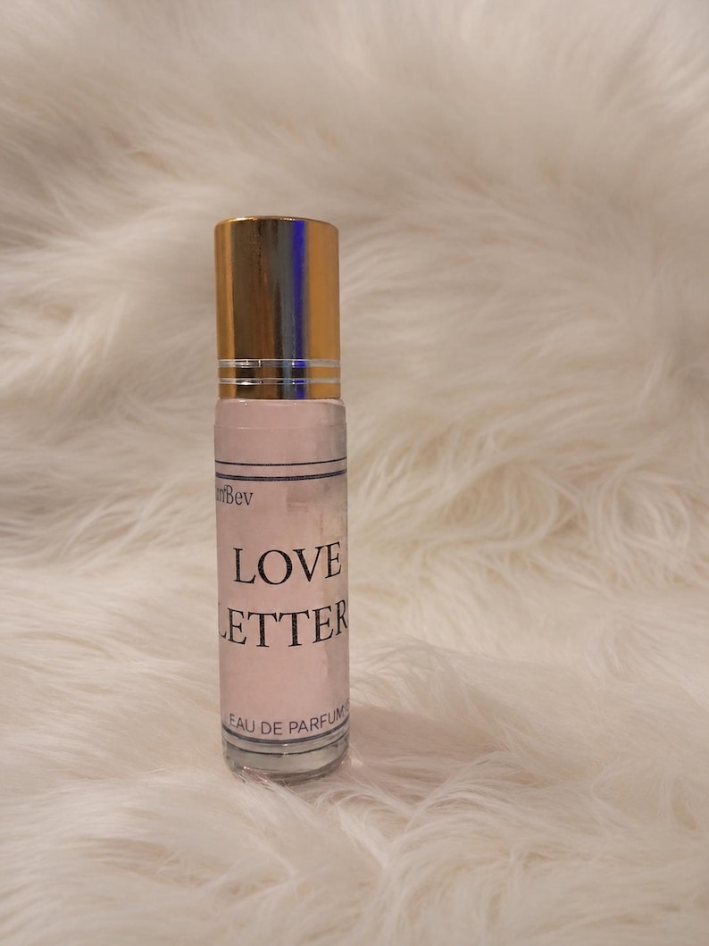 Puede incluir: Un frasco de perfume cil&iacute;ndrico de vidrio con una tapa de metal dorado. La etiqueta es de color rosa claro con texto negro que dice Love Letter y Eau de Parfum. El frasco contiene un l&iacute;quido transparente y tiene un aplicador de bola arriba.