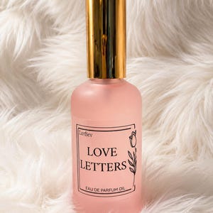 Puede incluir: Un frasco de perfume de vidrio rosa esmerilado con una tapa cil&iacute;ndrica dorada brillante. La etiqueta rectangular presenta un borde negro, una peque&ntilde;a ilustraci&oacute;n de un tulip&aacute;n y el texto LuvBev, LOVE LETTERS y EAU DE PARFUM OIL en negro.