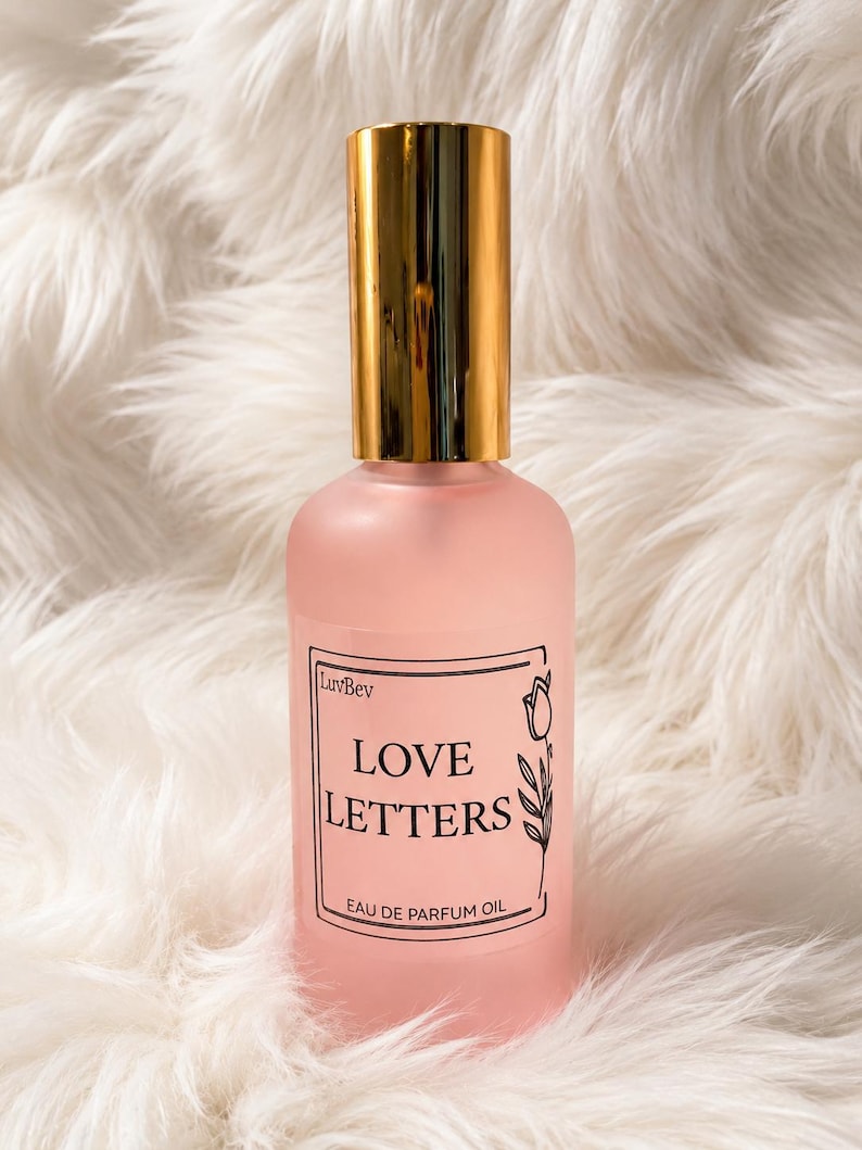 Puede incluir: Un frasco de perfume de vidrio rosa esmerilado con una tapa cil&iacute;ndrica dorada brillante. La etiqueta rectangular presenta un borde negro, una peque&ntilde;a ilustraci&oacute;n de un tulip&aacute;n y el texto LuvBev, LOVE LETTERS y EAU DE PARFUM OIL en negro.