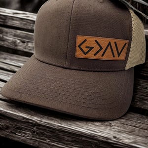 May include: A brown trucker hat with a mesh back and a curved brim. The hat features a rectangular patch with the letters "G>AV" in black. The hat is sitting on a wooden surface.