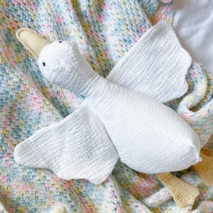 May include: A white stuffed goose toy with a beige beak and feet rests on a pastel-colored crocheted blanket. The goose has black eyes and textured wings. The blanket features a mix of pink, blue, and yellow yarn.