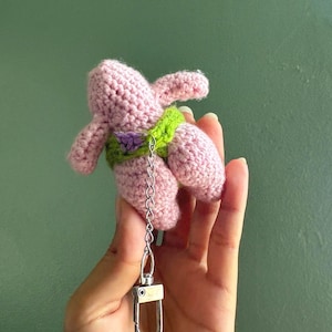 May include: A pink crocheted plush figure with a light green waistband and a small purple accent, attached to a silver-toned metal chain with a lobster clasp. The soft, textured yarn construction forms a character with limbs and a rounded body shape.