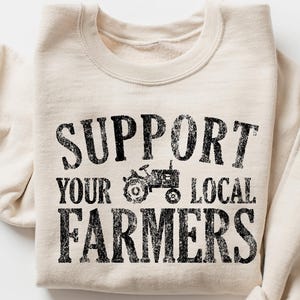 May include: Cream-colored sweatshirt with the text "SUPPORT YOUR LOCAL FARMERS" in distressed black lettering. A tractor graphic is centered above the word "FARMERS". The sleeves are folded, and the sweatshirt is on a white background.