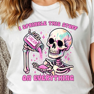 May include: White t-shirt with a colorful skeleton graphic. The skeleton is pouring pink glitter from a container labeled "Sarcasm." The text "I Sprinkle This Stuff On Everything" is above and below the graphic.