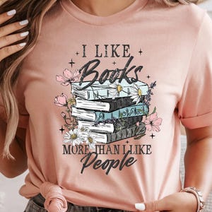 May include: A light pink t-shirt with the text "I Like Books More Than I Like People" in a whimsical font. The graphic features a stack of books adorned with flowers. The shirt has short sleeves and a casual fit.