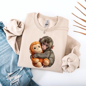May include: A beige sweatshirt featuring a graphic of two monkeys hugging. One monkey is gray, and the other is orange. The sweatshirt is paired with blue jeans with distressed details.