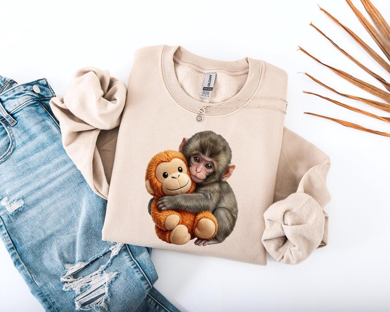 May include: A beige sweatshirt featuring a graphic of two monkeys hugging. One monkey is gray, and the other is orange. The sweatshirt is paired with blue jeans with distressed details.