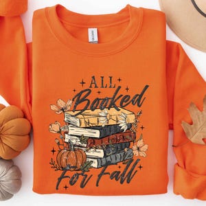 May include: An orange crewneck sweatshirt featuring a graphic of a stack of four books and a pumpkin surrounded by flowers. The text reads ALL Booked For Fall in a black distressed font. The design includes small star accents throughout the graphic.