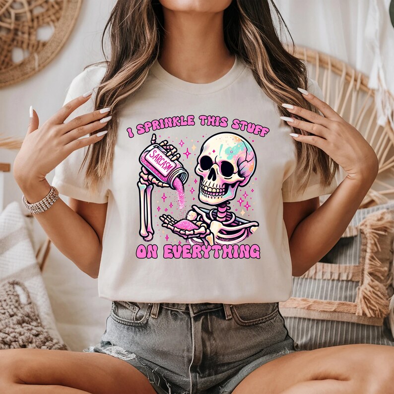 May include: Cream-colored t-shirt featuring a colorful skeleton graphic. The skeleton is holding a container labeled "Sarcasm" and sprinkling it onto its hand. The text "I Sprinkle This Stuff On Everything" is printed on the shirt.