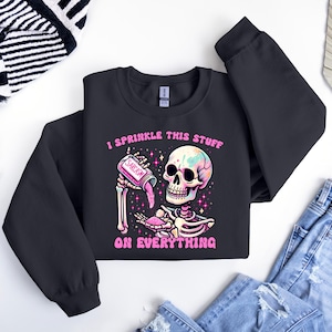 May include: Black sweatshirt with a colorful skeleton graphic. The skeleton is holding a jar labeled "Sarcasm" and sprinkling it. The text "I sprinkle this stuff on everything" is above and below the graphic. The sweatshirt is paired with blue jeans.