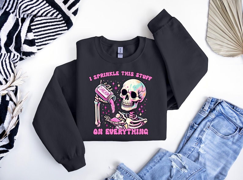 May include: Black sweatshirt with a colorful skeleton graphic. The skeleton is holding a jar labeled "Sarcasm" and sprinkling it. The text "I sprinkle this stuff on everything" is above and below the graphic. The sweatshirt is paired with blue jeans.