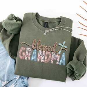May include: Olive green sweatshirt with the words "blessed grandma" in a decorative font. The word "grandma" is in a floral design with butterflies. A silver necklace with a pendant is draped on the sweatshirt.
