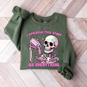 May include: An olive green sweatshirt featuring a colorful skeleton graphic. The skeleton is holding a jar labeled "Sarcasm" and sprinkling pink glitter. The text "I sprinkle this stuff on everything" is printed above and below the skeleton.