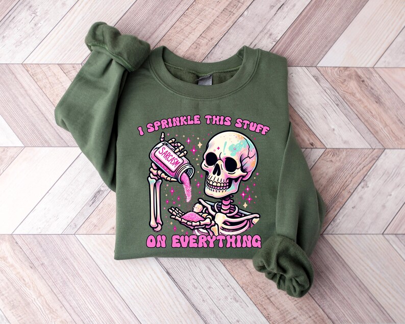 May include: An olive green sweatshirt featuring a colorful skeleton graphic. The skeleton is holding a jar labeled "Sarcasm" and sprinkling pink glitter. The text "I sprinkle this stuff on everything" is printed above and below the skeleton.