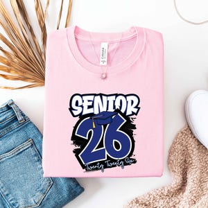 May include: A light pink t-shirt with the text "SENIOR 26 Twenty Twenty Six" in white and blue. The design includes a graduation cap graphic. The shirt is folded and placed on a white surface with blue jeans and white shoes.