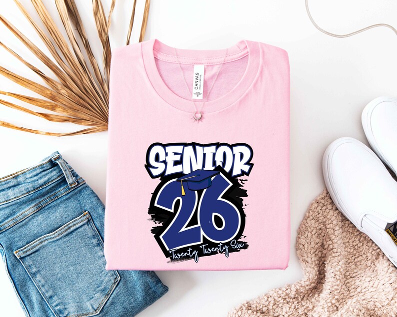 May include: A light pink t-shirt with the text "SENIOR 26 Twenty Twenty Six" in white and blue. The design includes a graduation cap graphic. The shirt is folded and placed on a white surface with blue jeans and white shoes.