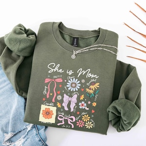 May include: Olive green sweatshirt with the text "She is Mom" and floral and bow graphics. A silver necklace with a pendant is draped over the sweatshirt. The sleeves are cuffed, and the sweatshirt is paired with blue jeans.